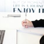 Close-up of a person's hand writing notes in an office setting with a laptop.