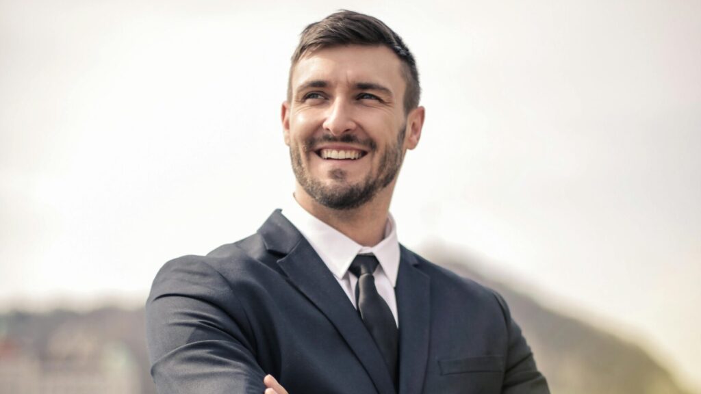 Smiling businessman in a black suit standing outdoors by a railing, showcasing a confident demeanor.