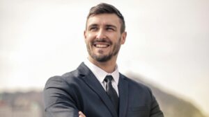 Smiling businessman in a black suit standing outdoors by a railing, showcasing a confident demeanor.