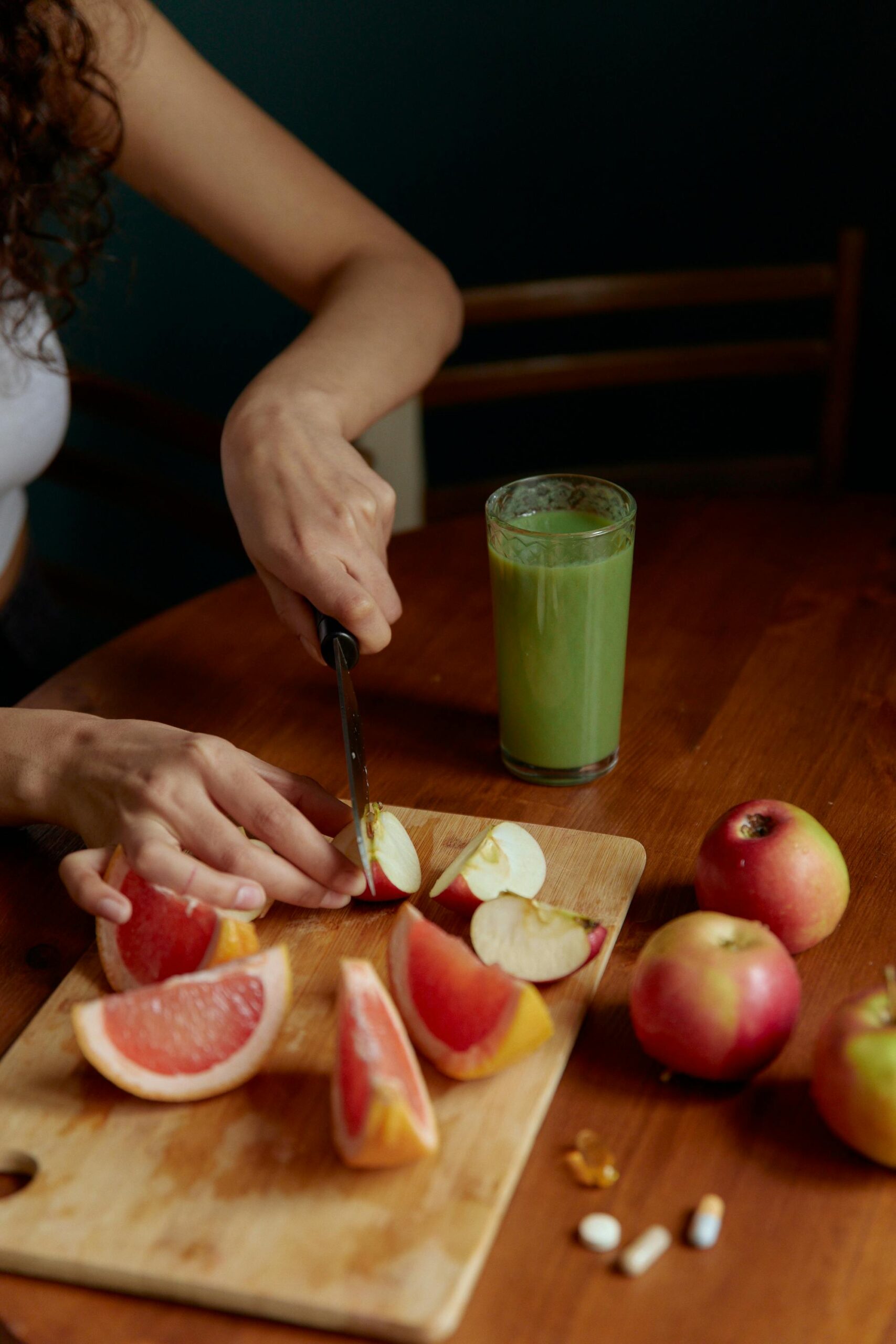Person slicing fruits with a knife, next to a green juice and medicine on a wooden board.