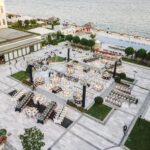 Aerial view of a lavish outdoor event setting near a waterfront, showcasing elegant tables and decorations.
