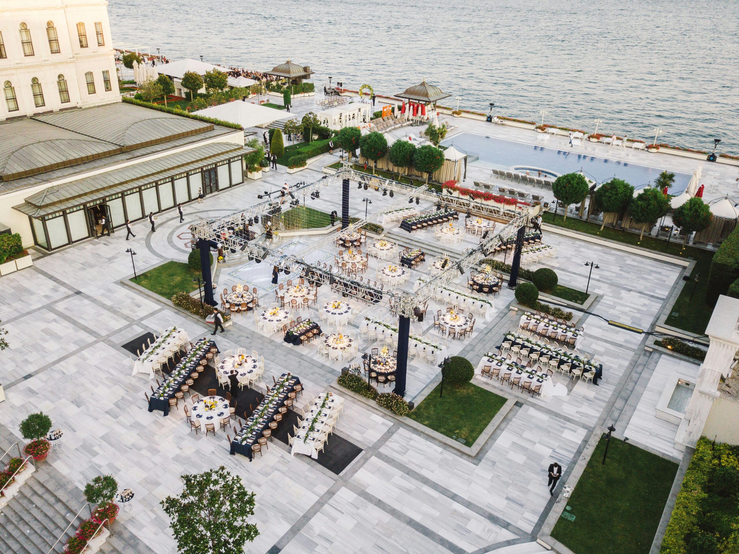 Aerial view of a lavish outdoor event setting near a waterfront, showcasing elegant tables and decorations.